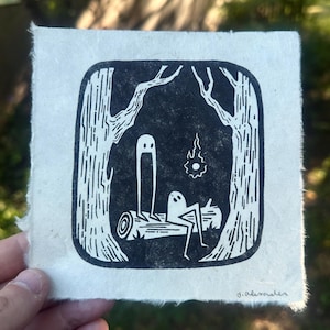 May include: A black and white linocut print depicting two ghost-like figures lounging on a log in a forest scene. The print includes a stylized flame and trees. The artist's signature, "d.alexander", is in the lower right corner.