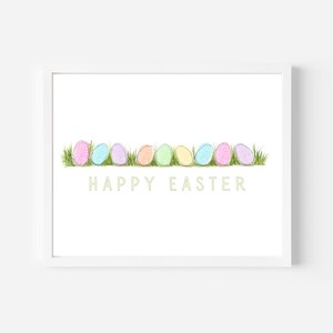 May include: A white framed print featuring a row of pastel coloured Easter eggs on a green grass background. The text "HAPPY EASTER" is written in a light green font below the eggs.
