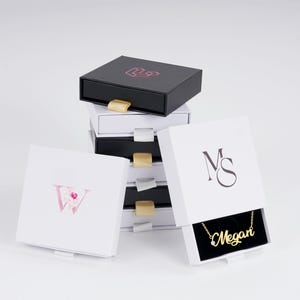 May include: A stack of jewelry boxes in black and white, some open to reveal a gold necklace. The boxes have gold and silver accents. One box has the initial "W" in pink floral design, and another has the initials "MS". The necklace says "Megan".