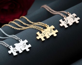 May include: Three gold, silver and rose gold necklaces with matching puzzle piece pendants. Each pendant has two pieces with initials engraved on them. The necklaces are on a black surface.