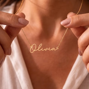 May include: A gold-toned name necklace featuring the name Olivia written in a cursive script font. The pendant is attached to a delicate, thin gold-toned chain that rests around the neck, showcasing a polished metallic finish and a simple design.