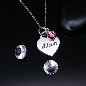 May include: Silver heart-shaped pendant necklace with the name "Alison" engraved on it. The pendant has a small pink crystal attached to it.