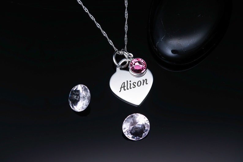 May include: Silver heart-shaped pendant necklace with the name "Alison" engraved on it. The pendant has a small pink crystal attached to it.