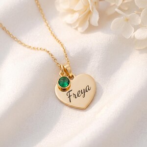 以下が含まれることがあります： 筆記体で「Freya」と刻印されたハート型のペンダントが付いたゴールドトーンのネックレス。ペンダントの上部には、ベゼルセットされた小さな丸いエメラルドグリーンの宝石があしらわれており、繊細なチェーンから吊り下げられています。