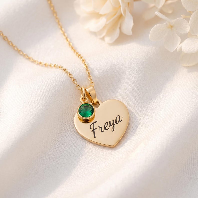 以下が含まれることがあります： 筆記体で「Freya」と刻印されたハート型のペンダントが付いたゴールドトーンのネックレス。ペンダントの上部には、ベゼルセットされた小さな丸いエメラルドグリーンの宝石があしらわれており、繊細なチェーンから吊り下げられています。