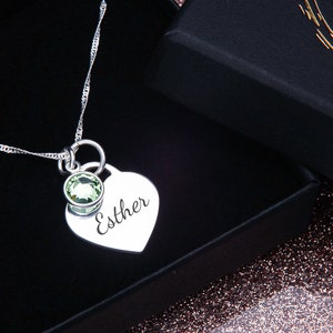 May include: A silver necklace with a heart-shaped pendant engraved with the name "Esther". The pendant has a small green gemstone attached to it.