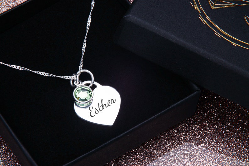 May include: A silver necklace with a heart-shaped pendant engraved with the name "Esther". The pendant has a small green gemstone attached to it.