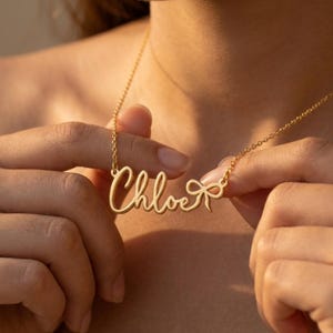 May include: A gold-toned necklace featuring a cursive nameplate pendant that reads Chloe, with a small decorative bow detail attached to the end of the name. The pendant is suspended from a delicate gold-toned chain that rests against the skin.
