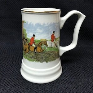 May include: White ceramic beer stein with a gold rim and a scene of two men on horseback riding through a field. The men are wearing red jackets and black hats.