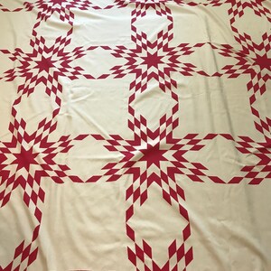 May include: A large, cream-colored quilt with a repeating pattern of red starbursts and diamond shapes. The quilt is spread out, showcasing the intricate design and craftsmanship. The overall theme is home decor.