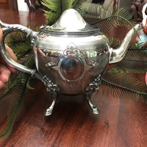 May include: A polished silver teapot featuring a rounded body with intricate engraved cross-hatching patterns, a central circular medallion with an engraved letter S, a curved spout, a domed lid, and three ornate legs with decorative floral motifs.