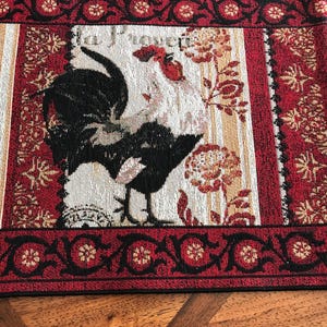 May include: Red, black, and white woven table runner with a rooster design and the text "La Poulette". The runner has a black and gold border with a floral pattern.