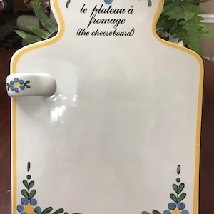 May include: White ceramic cheese board with a yellow border and blue and green floral designs. The board has a handle and two pineapple-shaped metal holders. The text "le plateau à fromage (the cheeseboard)" is written on the board.