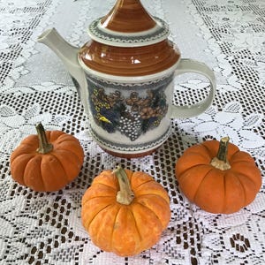 May include: A brown and white ceramic teapot with a floral design and a lid. Three small orange pumpkins are on a white lace tablecloth.