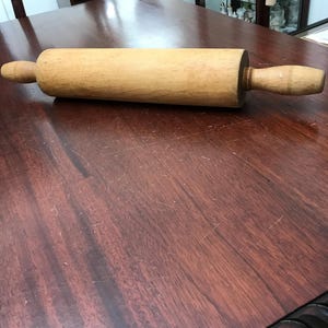 May include: A cylindrical wooden rolling pin with a smooth, light brown finish and two rounded handles on either end. The rolling pin features a central barrel section and tapered handles, showing natural wood grain patterns throughout the surface.