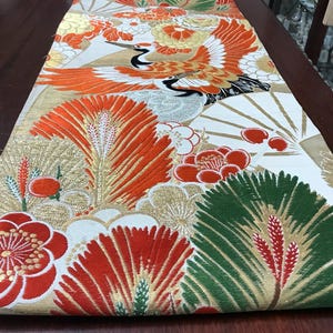 May include: An ornate table runner featuring a Japanese-inspired design. The runner showcases a cream background with gold accents and vibrant illustrations of cranes, fans, and floral patterns in red, orange, and green. The runner is approximately 72 inches long.