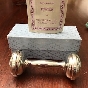 May include: A silver-colored dumbbell rattle made of pewter, with rounded ends and a central handle. The rattle is presented on a light blue box with a star pattern. A card with the text "Early American PEWTER" is placed on top.