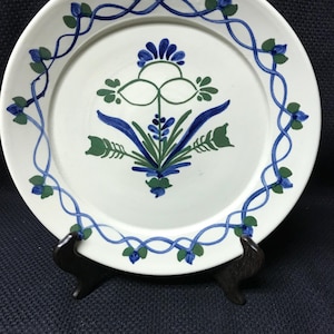 May include: White ceramic plate with a floral design in blue and green. The plate features a central floral motif and a border of intertwined vines and flowers. The plate is displayed on a dark brown stand.