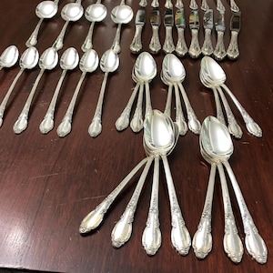 May include: A collection of ornate silver-colored flatware, including spoons, forks, and knives, arranged on a dark wooden surface. The silverware features detailed floral patterns on the handles, suggesting a vintage or antique style. The arrangement showcases a complete set.