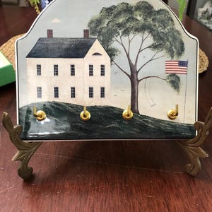 May include: A decorative key holder featuring a painted scene of a white house, a large tree, and an American flag. The key holder has four brass hooks and is supported by ornate metal legs.