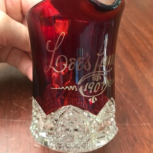 May include: A ruby red glass creamer with a clear, diamond-patterned base. The upper portion features a wavy rim and an etched inscription that reads "Lee's Summit" above the year "1907" enclosed in a decorative oval flourish.