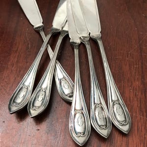May include: Set of six silver butter knives. Each knife features a wide, rounded blade and a long, slender handle. The handles have an engraved oval design with a script initial. The knives are arranged on a dark wooden surface.