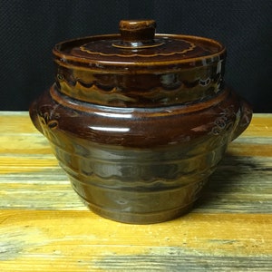May include: A brown ceramic crock with a lid. The crock has a wavy design and a brown glaze. The lid has a knob on top.