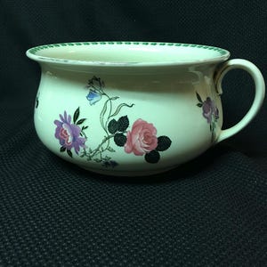 May include: A white ceramic chamber pot with a green rim and floral design featuring pink, purple, and blue flowers. The pot has a single handle.