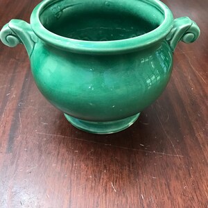 May include: A glossy, emerald green ceramic planter with a rounded body, a wide circular opening, and two decorative scroll-shaped handles on opposite sides. The vessel sits on a small, raised circular base and features a smooth, reflective finish.
