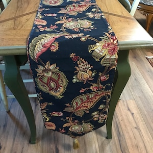 May include: A navy blue table runner with a floral pattern in shades of red, green, and gold. The runner has a tassel at the end.