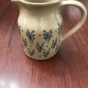 May include: A beige ceramic pitcher featuring a repeating dark blue hand-painted pattern of small sprigs and dots. The pitcher has a curved handle on the right side and a wide pouring spout at the top, with a smooth, glossy finish throughout the body.