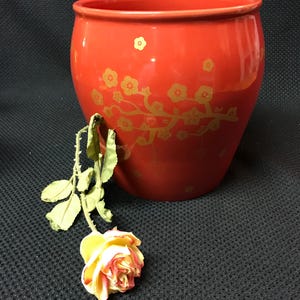 May include: A red ceramic planter with a gold floral design. The planter is sitting on a black surface with a yellow rose.