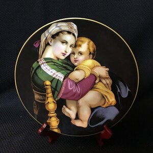 May include: A round porcelain plate with a black background and a gold rim. The plate features a painting of the Madonna and Child. The Madonna is wearing a green and purple robe with a white head covering. The child is wearing a yellow robe and is being held by the Madonna. The plate is on a wooden stand.