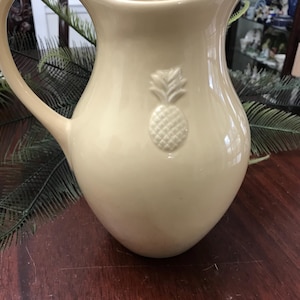 May include: A beige ceramic pitcher with a large handle and a pineapple emblem. The pitcher has a wide base and a curved spout. The pineapple is embossed on the front of the pitcher. The pitcher is sitting on a dark brown wooden surface.
