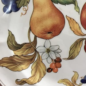 May include: A white ceramic plate with a hand-painted design of a pear, grapes, leaves, and flowers. The pear is yellow and orange, the grapes are purple, the leaves are green and brown, and the flowers are white with yellow centers.