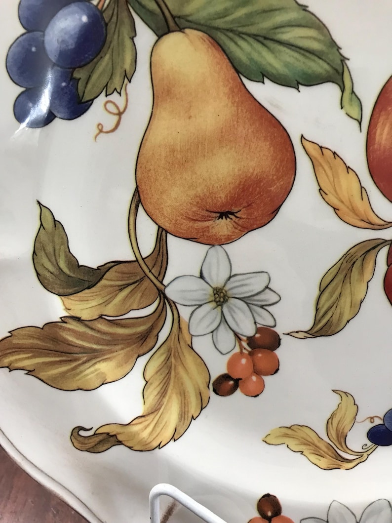 May include: A white ceramic plate with a hand-painted design of a pear, grapes, leaves, and flowers. The pear is yellow and orange, the grapes are purple, the leaves are green and brown, and the flowers are white with yellow centers.