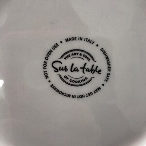 May include: Close-up of a white ceramic dish with a black label that reads "Sur la Table, The Art & Soul of Cooking." The label also states "Made in Italy, Dishwasher Safe, May Get Hot in Microwave, Not for Oven Use."