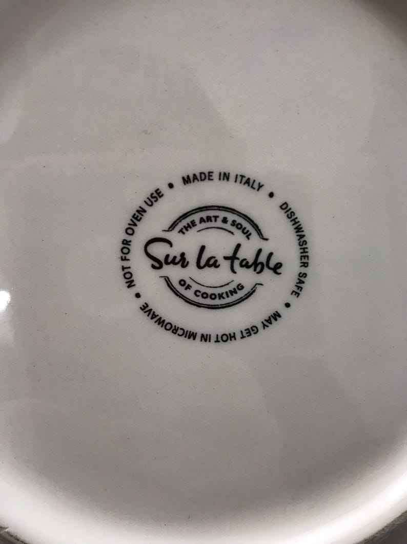 May include: Close-up of a white ceramic dish with a black label that reads "Sur la Table, The Art & Soul of Cooking." The label also states "Made in Italy, Dishwasher Safe, May Get Hot in Microwave, Not for Oven Use."