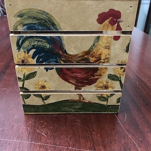 May include: A wooden box with a rooster and sunflower design. The rooster is painted with red, blue, yellow, and brown colors. The box is rectangular and has a light beige background.