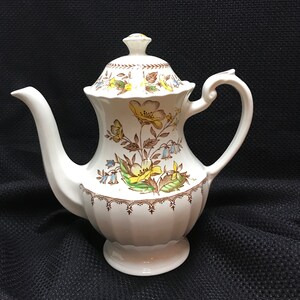 May include: A white ceramic coffee pot with a floral design in shades of brown, yellow, and blue. The pot has a lid and a handle.
