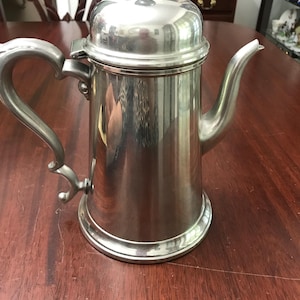 May include: A silver-colored, vintage coffee pot with a lid and a curved handle. The pot has a spout and sits on a round base. The pot is made of metal and reflects the surrounding environment.