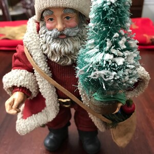 May include: A detailed figurine of Santa Claus, dressed in a red suit with white trim and a matching hat. He carries a small green Christmas tree and a tan satchel. The figurine has a detailed face and is a festive holiday decoration.