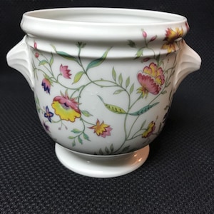 May include: White ceramic planter with a floral design. The planter features a pedestal base, two side handles, and a wide rim. The floral pattern includes pink, yellow, and purple flowers with green leaves and vines. The planter is suitable for indoor or outdoor use.