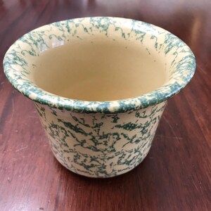 May include: A cream-colored ceramic bowl with a green splatter pattern. The bowl has a wide, slightly flared rim and a rounded body. The interior is a solid cream color. The bowl is likely vintage or antique.