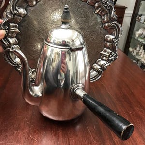 May include: A silver-colored teapot with a black handle and a decorative lid, set against a silver tray with an ornate, embossed design. The tray has a detailed, scrolled border, and the teapot reflects the surrounding environment.