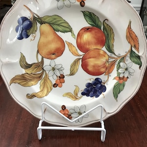May include: A white ceramic plate with a decorative design of fruit and flowers. The plate features pears, apples, grapes, and white flowers with red berries. The plate is on a white stand.