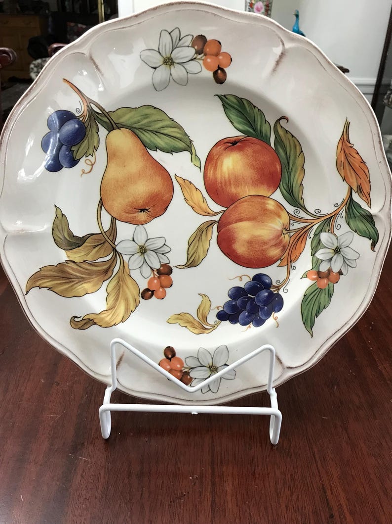 May include: A white ceramic plate with a decorative design of fruit and flowers. The plate features pears, apples, grapes, and white flowers with red berries. The plate is on a white stand.
