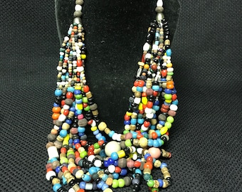May include: Multi-strand necklace made of colorful glass and wooden beads. The necklace is layered and features a variety of colors, including red, blue, green, yellow, and white.