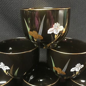 May include: Set of six black teacups with gold rims and a floral design featuring white and brown irises.