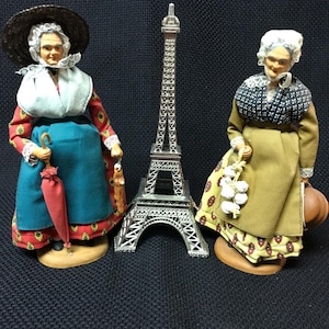 May include: Two vintage figurines of women in traditional attire, standing on wooden bases, flank a silver Eiffel Tower replica. One figure wears a hat and holds an umbrella, the other carries a basket and a jug. The background is dark.